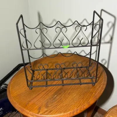 Sale Photo Thumbnail #326: Black wrought iron two-tier wine rack featuring decorative scrollwork detail, sitting on a round wooden end table.