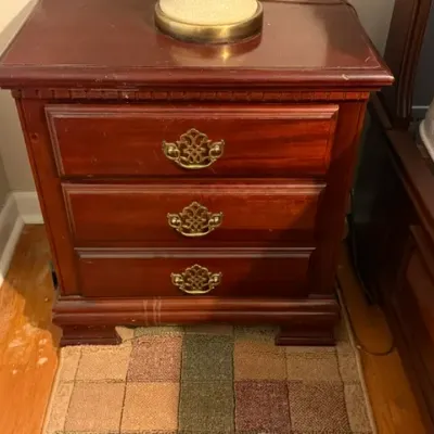 Sale Photo Thumbnail #159: Traditional cherry-finish wood nightstand with three drawers and ornate brass bale handles, showing light surface wear.