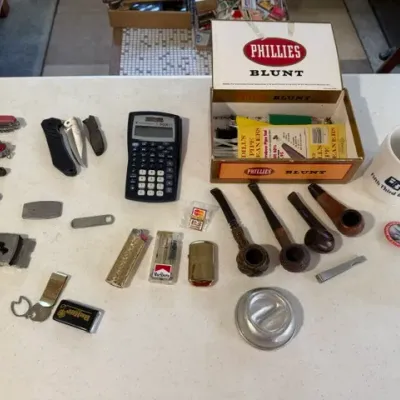 Sale Photo Thumbnail #224: Collection of vintage smoker's accessories and pocket items, including wooden tobacco pipes, pocket knives, lighters, pipe cleaners in a Phillies Blunt cigar box, and a scientific calculator.