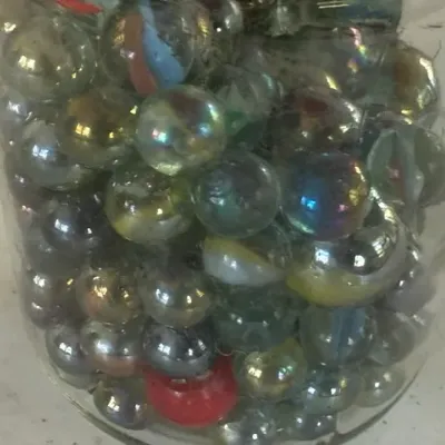 Sale Photo Thumbnail #245: Collection of vintage glass marbles, featuring a variety of iridescent finish, cat's-eye, and solid color designs stored in a clear glass jar.