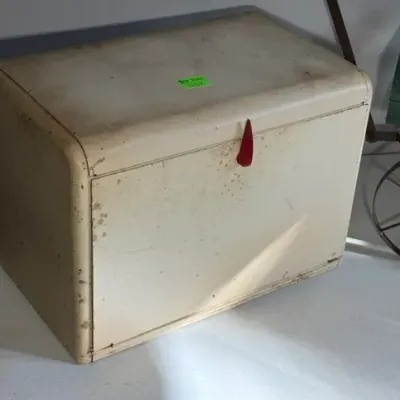 Sale Photo Thumbnail #437: Vintage off-white metal bread box featuring a red plastic latch handle and rounded edges, displaying age-related wear and surface scuffs.