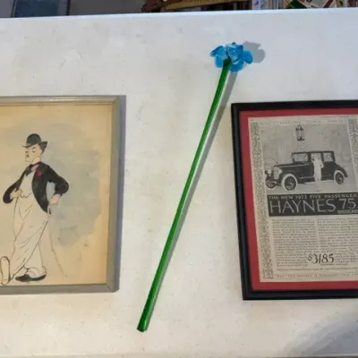 Sale Photo Thumbnail #220: Framed Charlie Chaplin-style illustration and a framed 1922 Haynes 75 vintage advertisement, alongside a decorative long-stemmed blue glass flower.