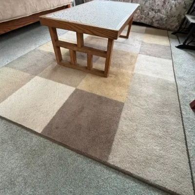 Sale Photo Thumbnail #184: Large area rug featuring a colorblock grid pattern in neutral beige, tan, and brown tones, positioned under a mid-century wooden coffee table with a tiled top.