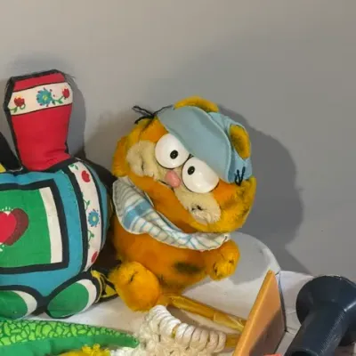 Sale Photo Thumbnail #435: Vintage Garfield plush wearing a nightcap alongside a colorful fabric train pillow and miscellaneous small items.