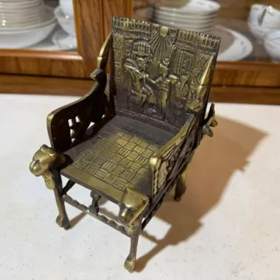 Sale Photo Thumbnail #109: Miniature brass and metal Egyptian Revival King Tut throne chair, featuring detailed relief carvings and lion head armrests.