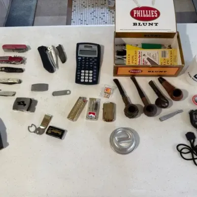 Sale Photo Thumbnail #223: Assortment of small collectibles and everyday items including pocket knives, smoking pipes, lighters, a Texas Instruments scientific calculator, and a vintage Phillies Blunt cigar box.
