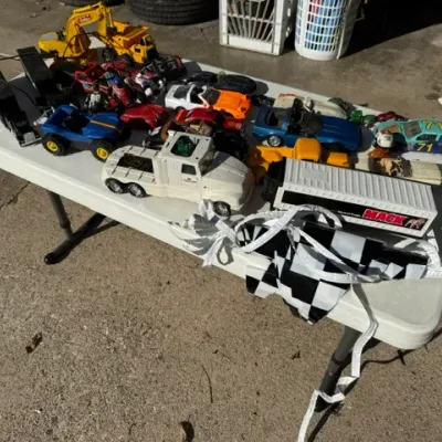 Sale Photo Thumbnail #467: Collection of vintage toy vehicles displayed on a white folding table, including a Tonka construction set, a Mack semi-truck, and various die-cast and plastic sports cars.