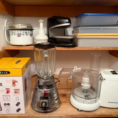 Sale Photo Thumbnail #273: Collection of small kitchen appliances on wooden shelves, including an Osterizer chrome blender, Hamilton Beach food processor, boxed Bella Rocket Blender, and a vintage floral slow cooker.