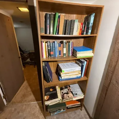Sale Photo Thumbnail #357: Tall wood-grain laminate bookcase filled with a varied selection of vintage and modern books, manuals, and loose papers.