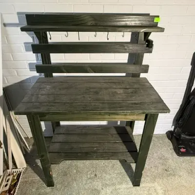 Sale Photo Thumbnail #198: Dark green stained wooden potting bench featuring an upper shelf with hanging hooks, a lower storage shelf, and slatted back construction.