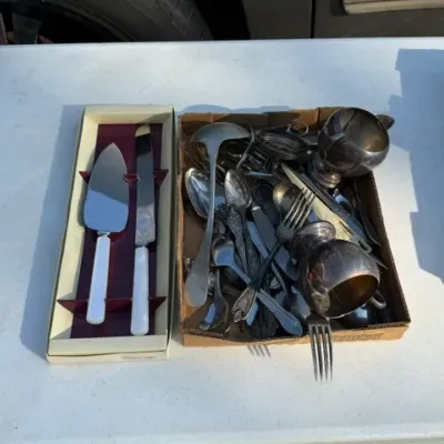 Sale Photo Thumbnail #447: Box of assorted silver-plated and metal flatware and serving pieces, alongside a boxed cake server set with white handles.