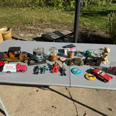 Sale Photo Thumbnail #469: Collection of vintage toy vehicles, action figures, snow globes, and novelty knick-knacks displayed on a outdoor folding table.
