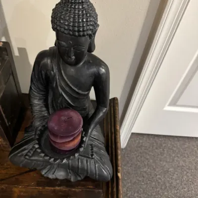 Sale Photo Thumbnail #28: Black seated Buddha candle holder statue holding a purple pillar candle, resting on a wooden side table.