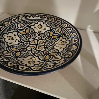 Sale Photo Thumbnail #41: Hand-painted Moroccan style ceramic plate featuring intricate geometric patterns in navy blue, yellow, and brown, displayed on a shelf.