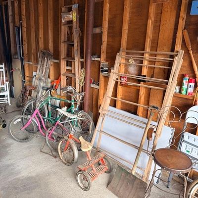 Collection of vintage garage items, including several bicycles, a child's tricycle, wooden ladders, and a wire-back bar stool.