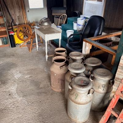 Collection of vintage metal milk cans alongside a wooden spinning wheel and small white painted table inside a sunroom.