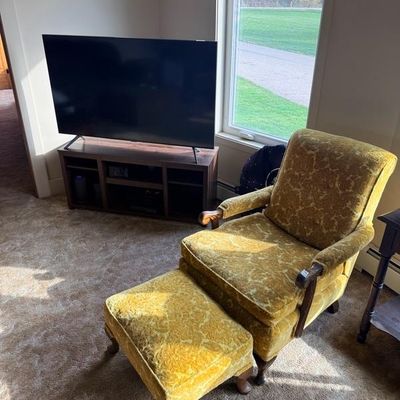 Vintage gold velvet armchair and matching ottoman with carved wood trim, alongside a modern flat-screen TV on a dark wood media console.