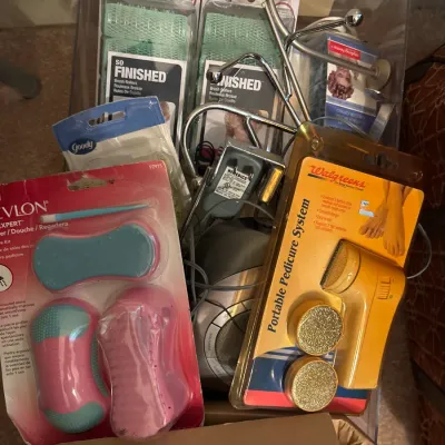 Assorted personal care items and small electronics in a clear plastic bin, including Goody brush rollers, a Revlon pedicure kit, and a...