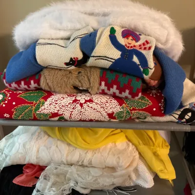 Stack of folded vintage clothing items, featuring novelty winter sweaters with embroidery, a white fur-trimmed coat, and lace garments.