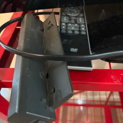 Black DVD player remote control alongside an open battery door or accessory part on a red metal wire shelf unit.