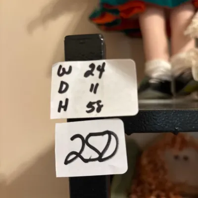 Black metal shelving unit holding dolls, labeled with dimensions of 24 inches wide, 11 inches deep, and 58 inches high, priced at 250.