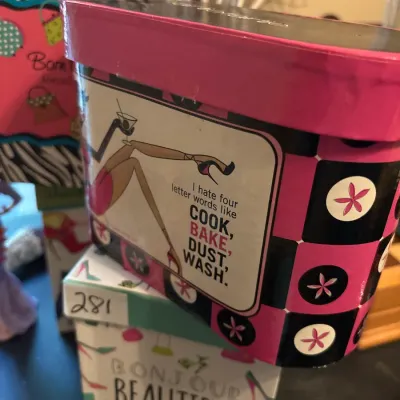 Novelty tin storage box featuring a humorous retro graphic and black and pink floral checker pattern, resting atop a decorative paper box.