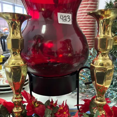 Ruby red glass hurricane lamp on a black metal stand, flanked by a pair of brass candlesticks and framed by artificial poinsettias.