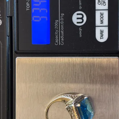 Gold-tone ring featuring a large emerald-cut blue stone and filigree detailing, shown on a digital scale displaying 9.34 grams.