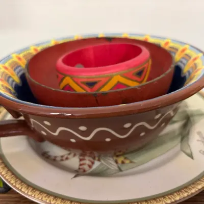 Hand-painted brown ceramic tea cup and small bowl nested on a botanical leaf saucer, paired with a brightly painted wooden bangle.