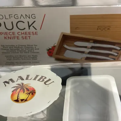 Wolfgang Puck 3-piece cheese knife set in original packaging, sitting behind a Malibu Rum branded item.
