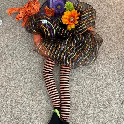 Decorative Halloween witch wreath featuring a pointed hat, artificial floral accents, metallic mesh mesh ribbon, and dangling striped legs.