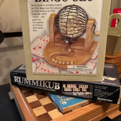 Stack of vintage board games including a Deluxe Solid Wood & Metal Bingo Set in its box, a Pressman Rummikub game, and a wooden...