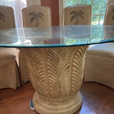 Tropical-style glass-top dining table featuring a carved palm leaf urn base, surrounded by matching palm tree embroidered slipcovered...