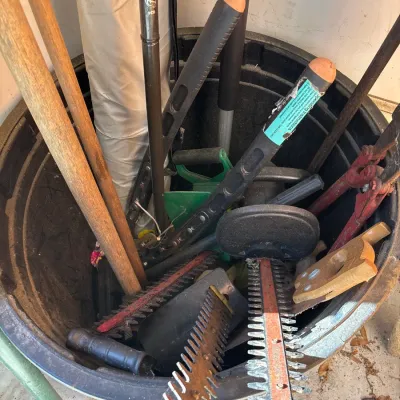 Black plastic trash bin filled with assorted yard tools, including electric hedge trimmers, wooden-handled implements, and bolt cutters.