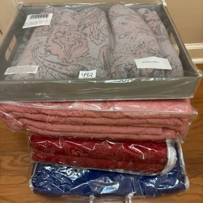 Stack of folded home textiles and linens, individually sealed in original plastic packaging, including a paisley set in a tray and...