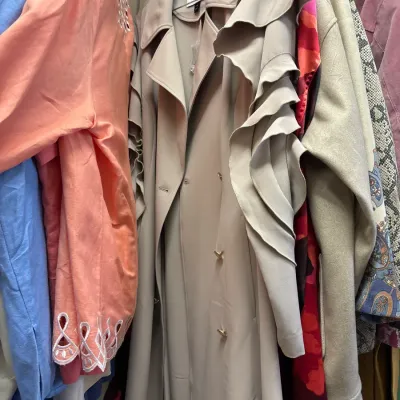 Tan trench coat featuring dramatic ruffled trim along the sleeves and shoulders, hanging among other colorful garments.