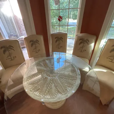 Tropical style dining set featuring a carved leaf stone-look pedestal table base with glass top, paired with five upholstered high-back...