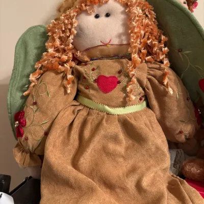 Handmade cloth angel doll featuring textured orange yarn hair, green felt wings, and an embroidered tan dress with a red heart motif.