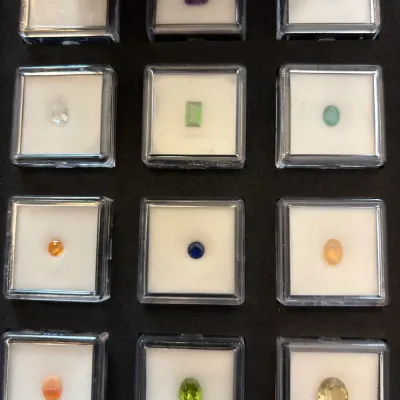 Collection of twelve loose faceted gemstones, including amethyst, sapphire, peridot, and citrine, displayed in individual square...