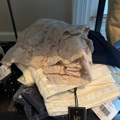Stack of women's apparel featuring cream-colored lace fabric, white folded denim, and dark garments, mostly with original retail...