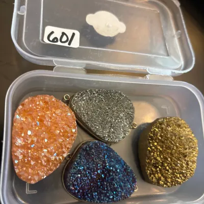 Set of four teardrop-shaped druzy crystal pendants in orange, silver, blue, and gold tones, stored in a small plastic organizer box.