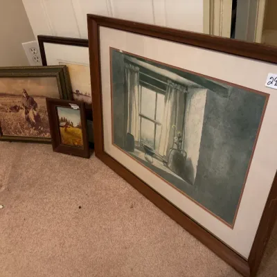 Collection of framed artwork leaned against a wall, including a large matted print of a window scene and various smaller landscape and...