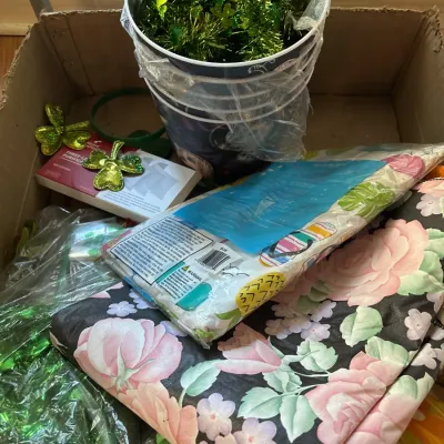 Cardboard box packed with St. Patrick's Day decorations, green tinsel, shamrock headbands, and floral patterned linens.