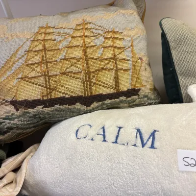 Vintage needlepoint throw pillow depicting a tall sailing ship at sea, shown alongside a white embroidered plush pillow.