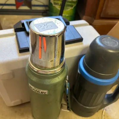 Vintage green Aladdin Stanley vacuum bottle alongside a black plastic thermos mug and a hard shell plastic carrying case.