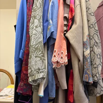 Closet rod filled with various women's garments, including patterned blouses, a light beige jacket with fringed trim, and a...