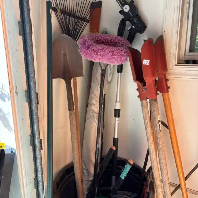 Collection of long-handled yard tools stored in a plastic tub, including shovels, rakes, post hole diggers, and a Sun Joe battery-powered...