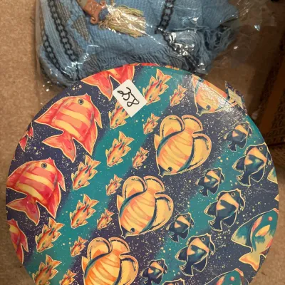 Round vintage decorative hat box featuring a colorful tropical fish motif alongside a wrapped light blue fringed shawl.