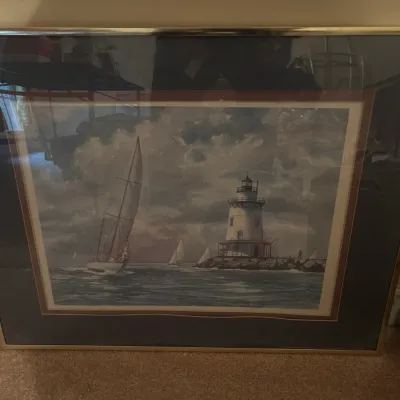 Framed nautical art print depicting a sailboat passing a lighthouse under a cloudy sky, presented in a double mat and silver-tone metal...