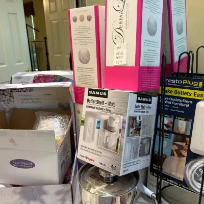 Collection of boxed household items including Sanus outlet shelves, a Presto Plug outlet extender, a FaceMaster facial toning system, and...
